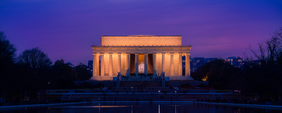 The Lincoln Memorial, illuminated from within with golden light. The sky behind it is purple, and there are dark trees to either side.